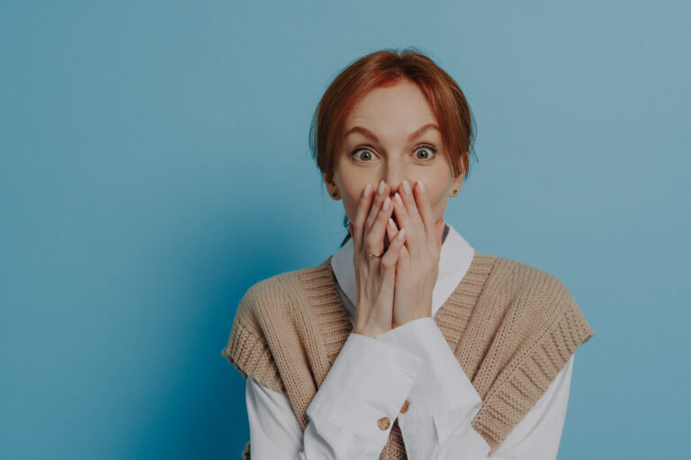 Shocked amazed redhead woman in stylish outfit covering mouth with both hands, isolated on blue